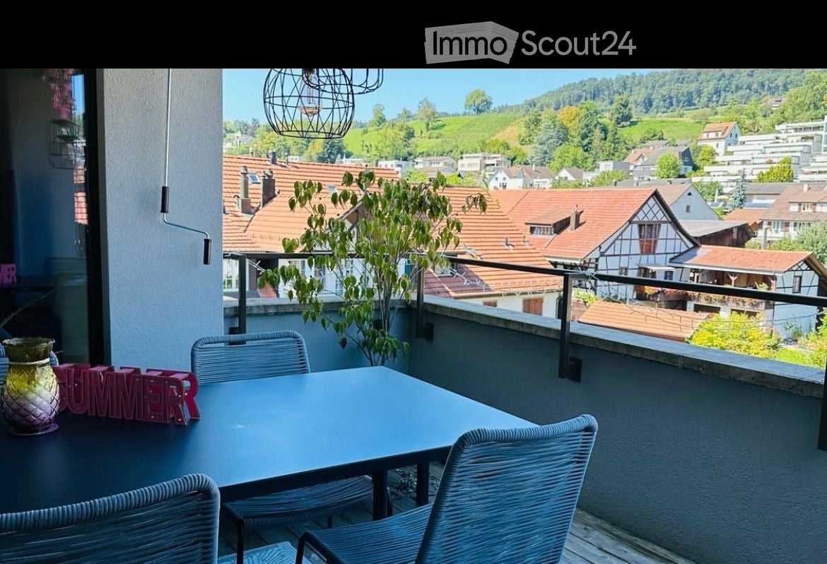 Balcony with a scenic view of a town and hills, featuring a table, chairs, and a decorative vase.