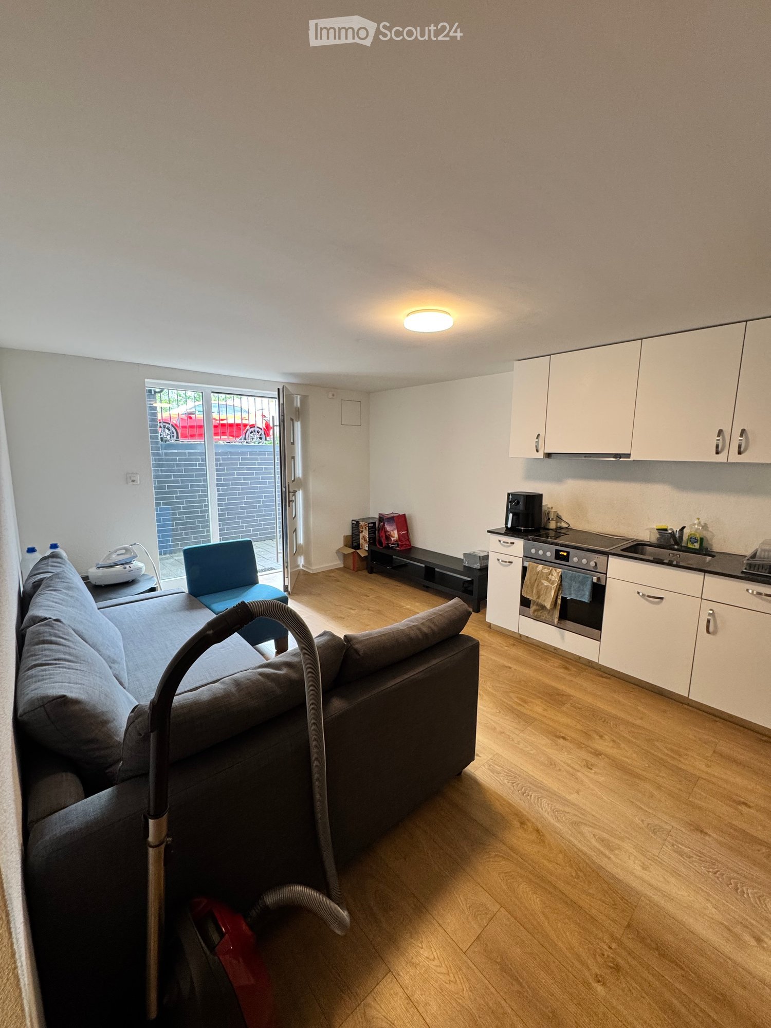 A modern, open-plan living and dining area with a grey sofa, a blue chair, a coffee table, and a small kitchen area with white cabinets, an oven, and a sink.