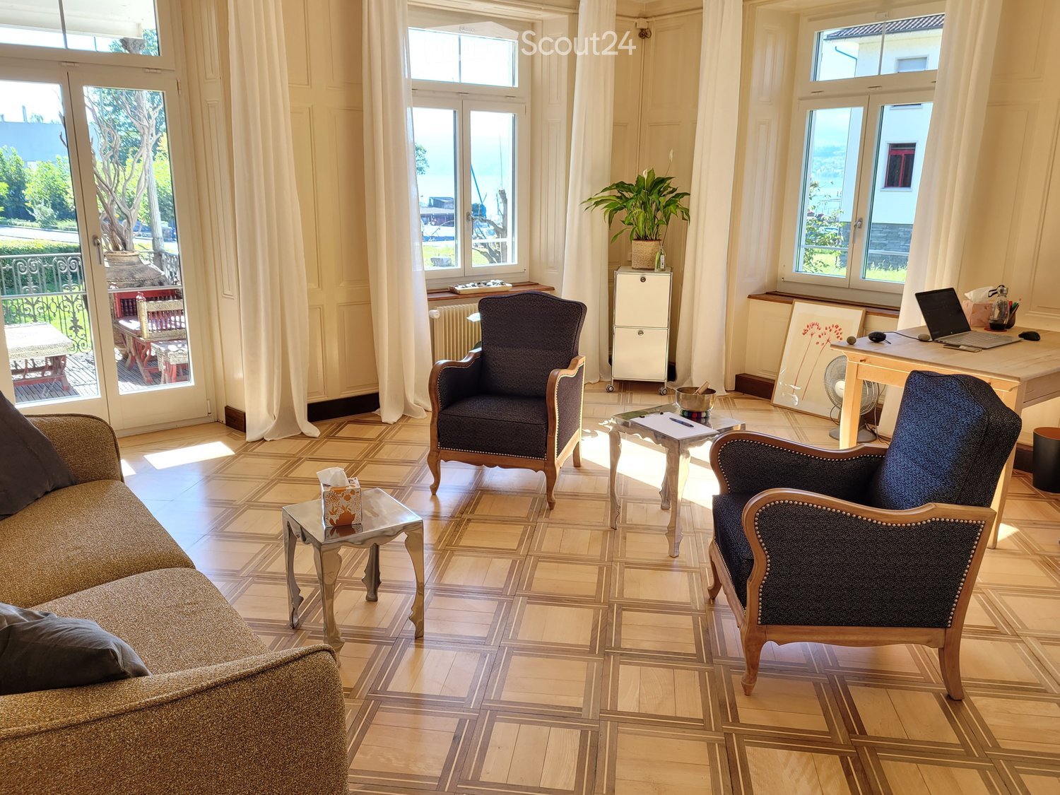 Bright living room with large windows and a view, featuring a sofa, two armchairs, a coffee table, a desk with a laptop, and a small plant on a side table.