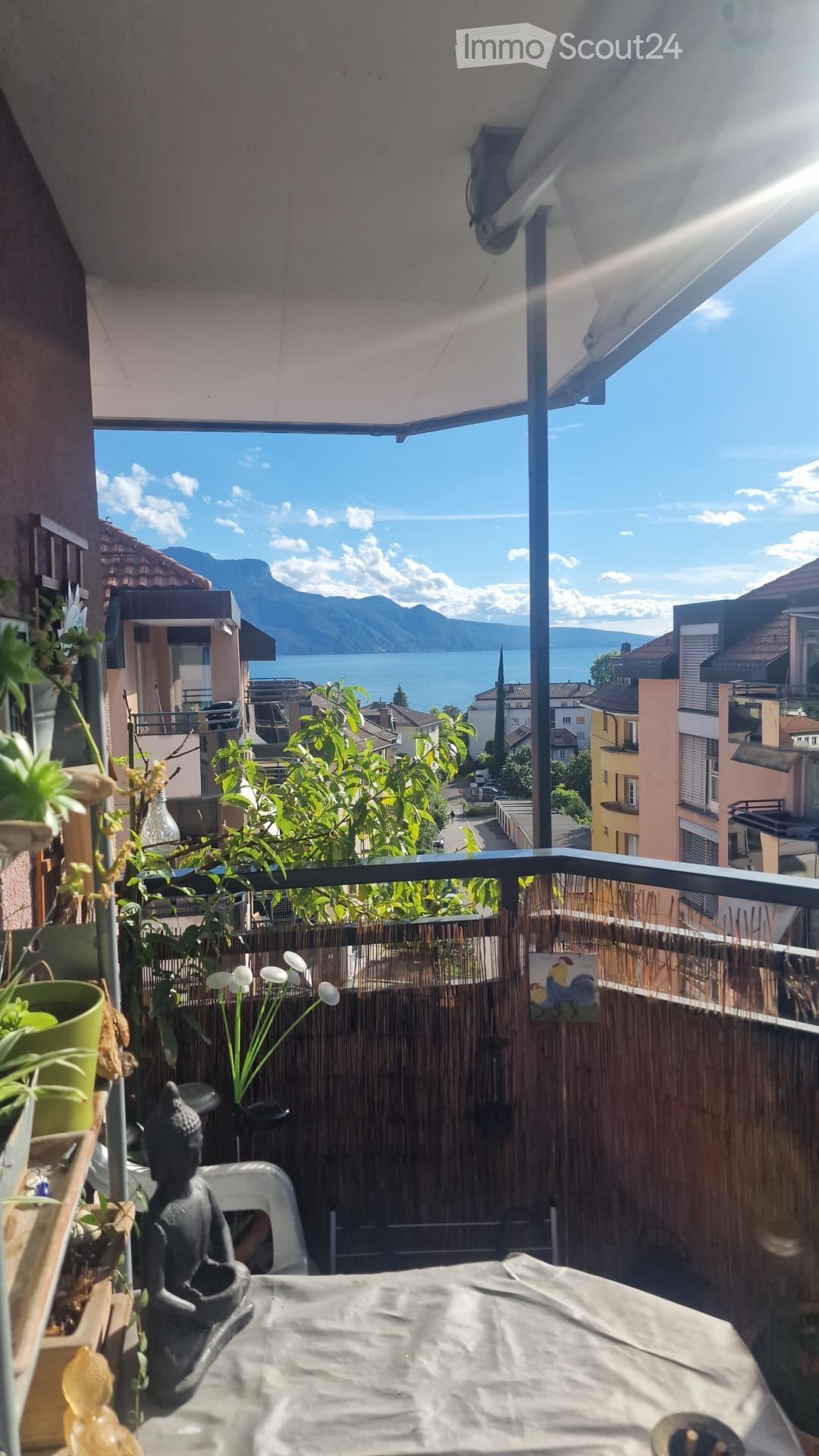 Balcony with a scenic view of a lake and mountains, featuring a table, chair, statue, plants, and an umbrella.