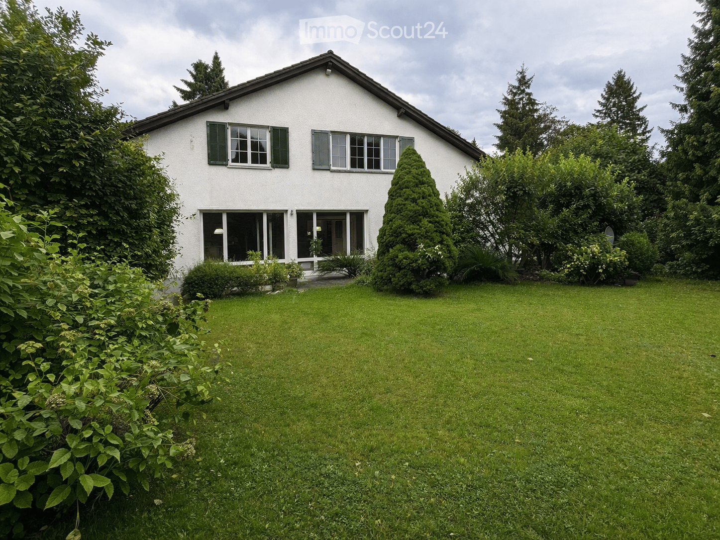 A two-story white house with green shutters and large windows, surrounded by a well-maintained lawn and trees.