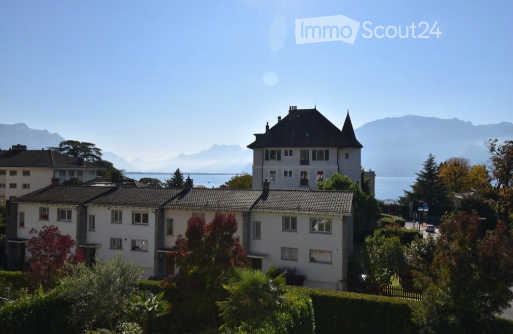 A multi-story residential building with a classic architectural style, surrounded by greenery and offering a scenic view of mountains and a body of water in the background.