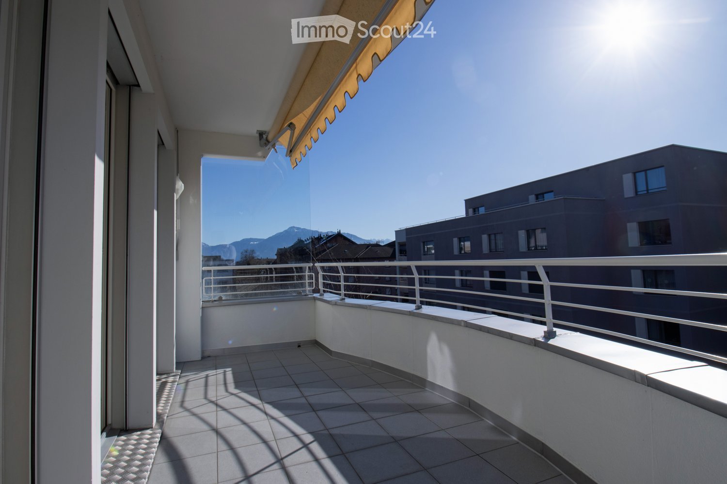 Balcony with tiled floor, white metal railing, and a retractable awning, overlooking a cityscape with buildings and mountains in the background.