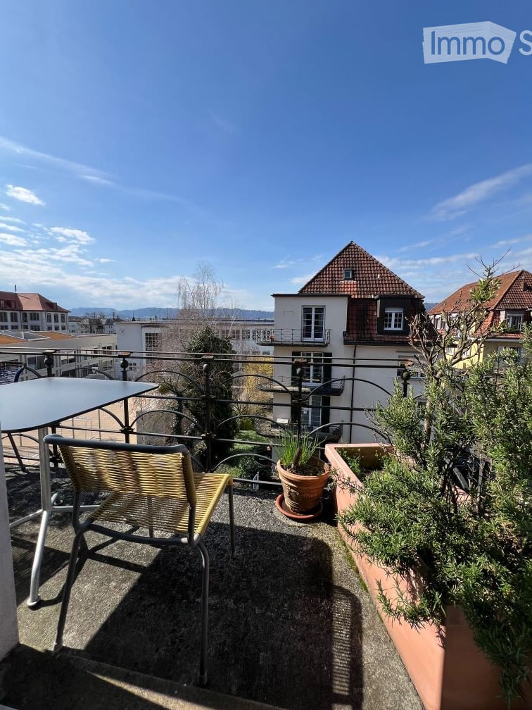 Balcony with a view of the city, featuring a metal railing, a wicker chair, a small table, and potted plants.