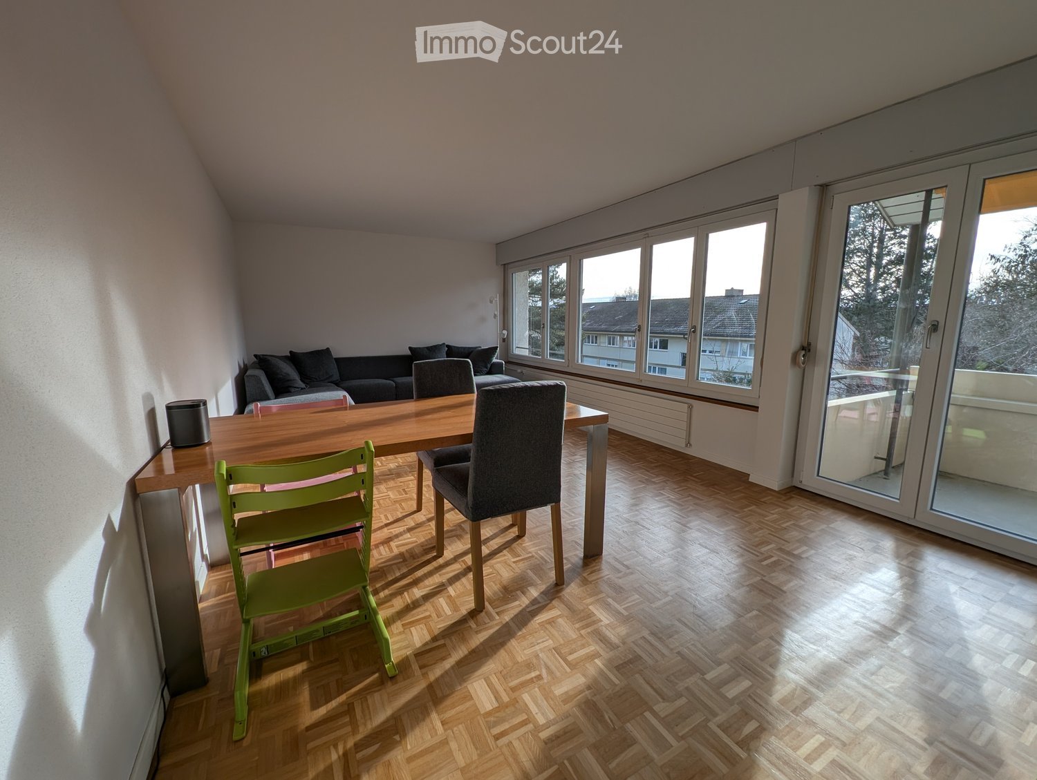 Bright living room with wooden herringbone flooring, large windows, and a sliding glass door leading to a balcony. Features a grey sofa, wooden dining table with green chairs, and a grey armchair.