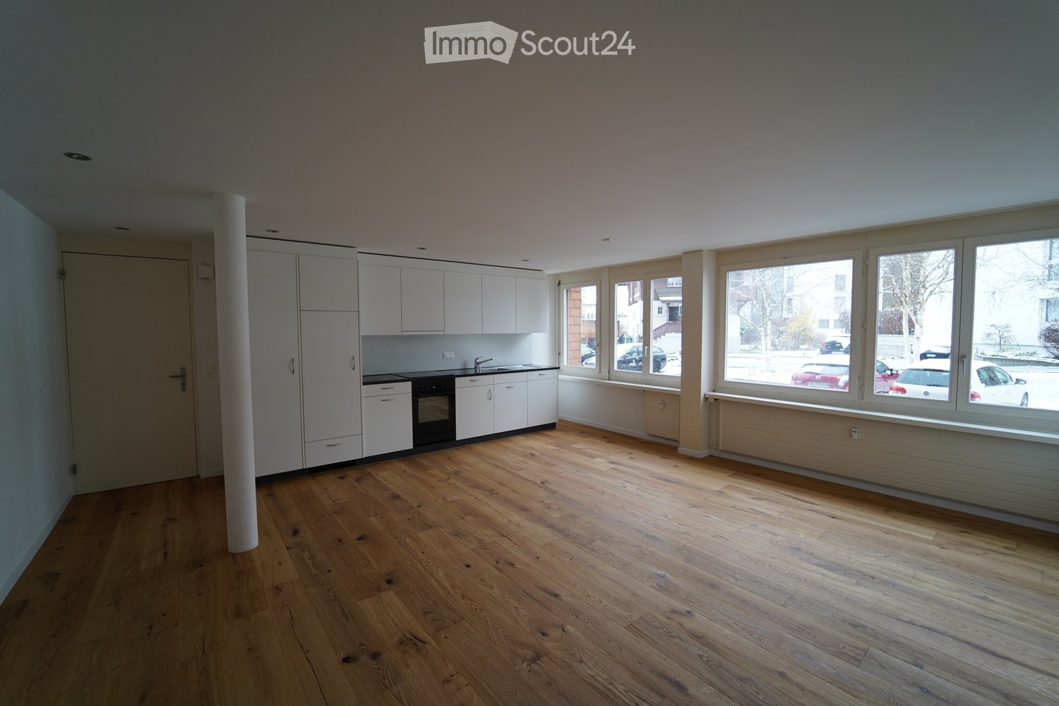 Empty room with light wooden flooring, white cabinetry, and large windows overlooking a street with parked cars.