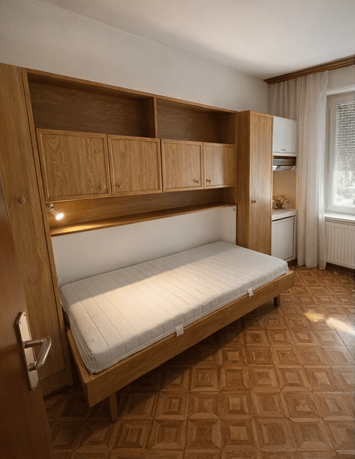 A compact bedroom with a single bed, built-in wooden wardrobe, and small cabinet. The room features wooden flooring and large windows with curtains.