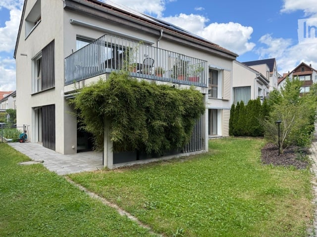 Modern two-story house with a balcony, green lawn, and surrounding trees.