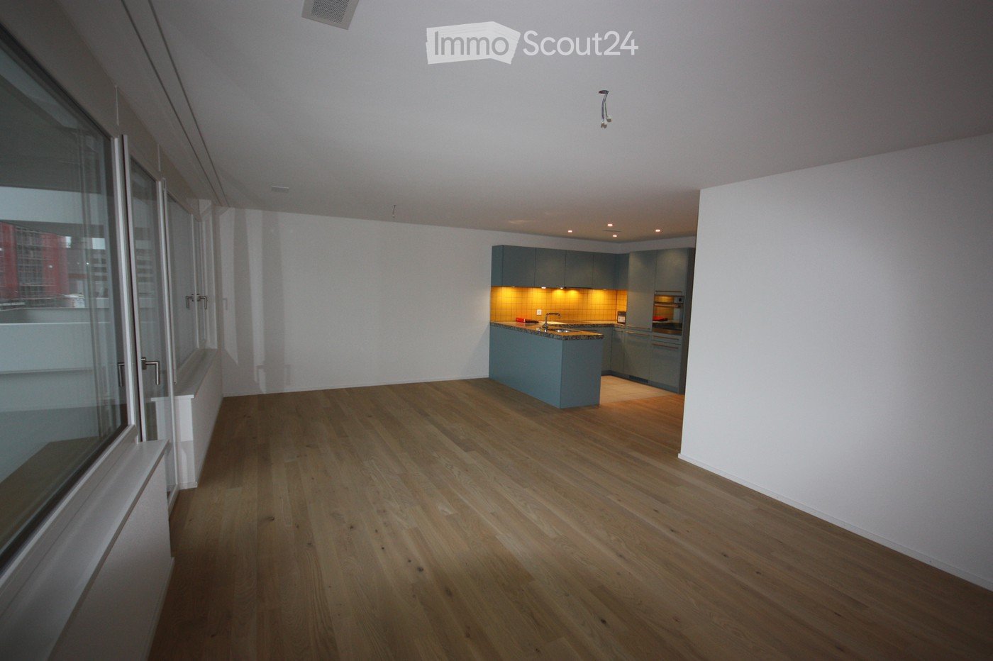 Open-plan space with wooden flooring, modern kitchen area featuring a kitchen island, cabinets, and a sink, large windows with a view, and recessed lighting.