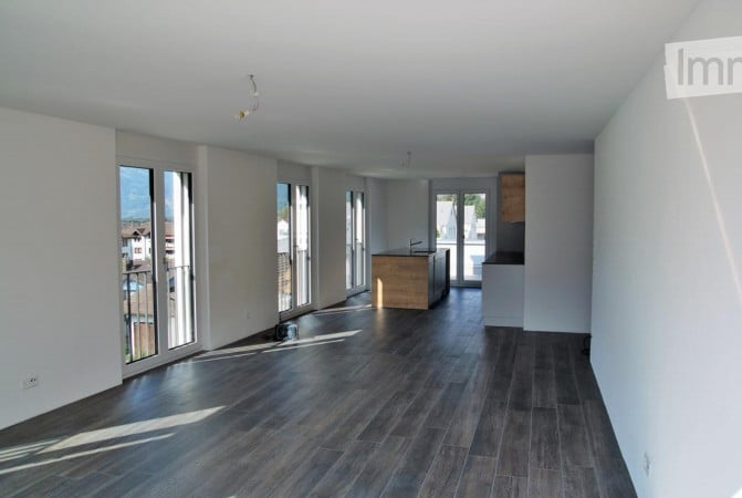 Empty room with dark wooden flooring, white walls, large windows, and a small kitchen area visible in the background.
