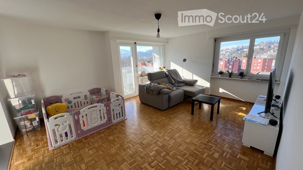 Bright living room with wooden flooring, large windows, and a view of the city and mountains. Features a grey sectional sofa, a small black coffee table, a TV stand, and a colorful playpen with a teddy bear.