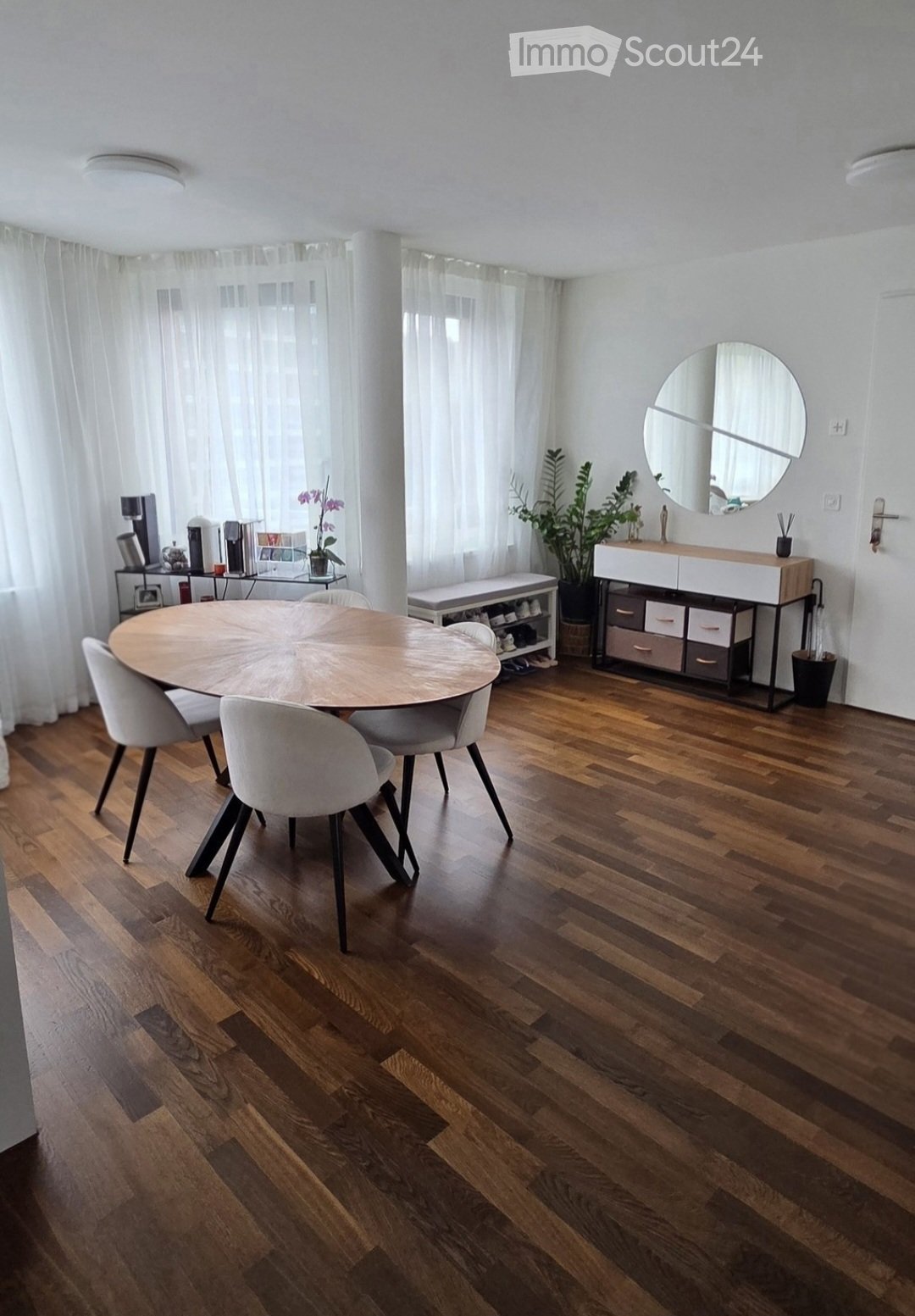 Bright dining area with wooden floor, round wooden table surrounded by white chairs, storage unit with drawers, bench, potted plants, and a round mirror on the wall.
