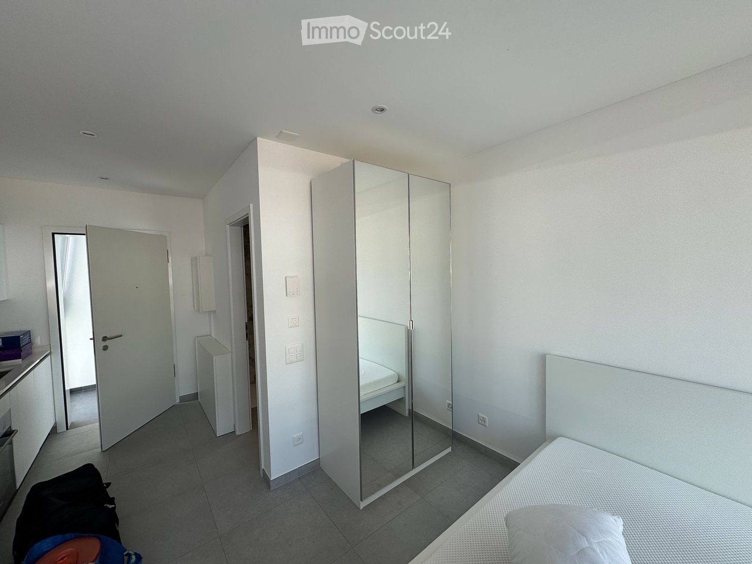 Modern, minimalist bedroom with a single bed, large mirrored wardrobe, and tiled floor. The room features white walls and a neutral color palette.