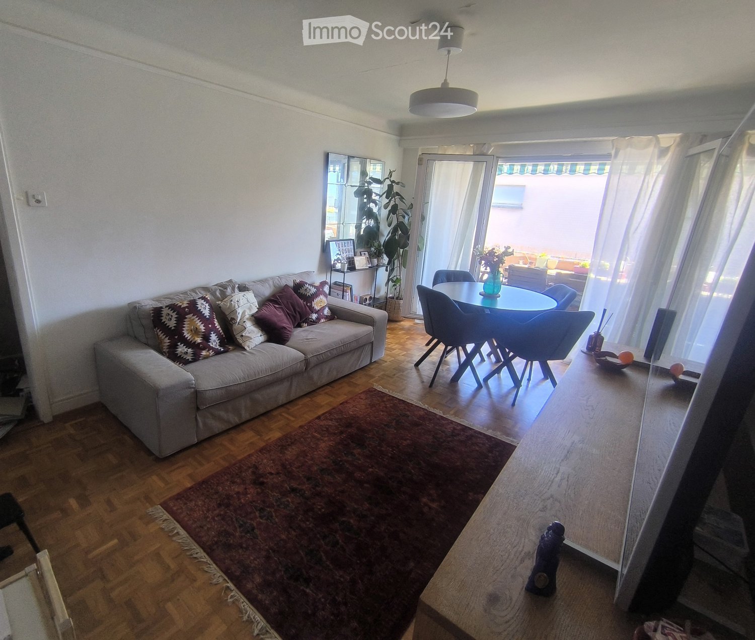 Bright living room with wooden flooring, a grey sofa with patterned cushions, a round dining table with chairs, a large mirror, and a potted plant near the balcony entrance.