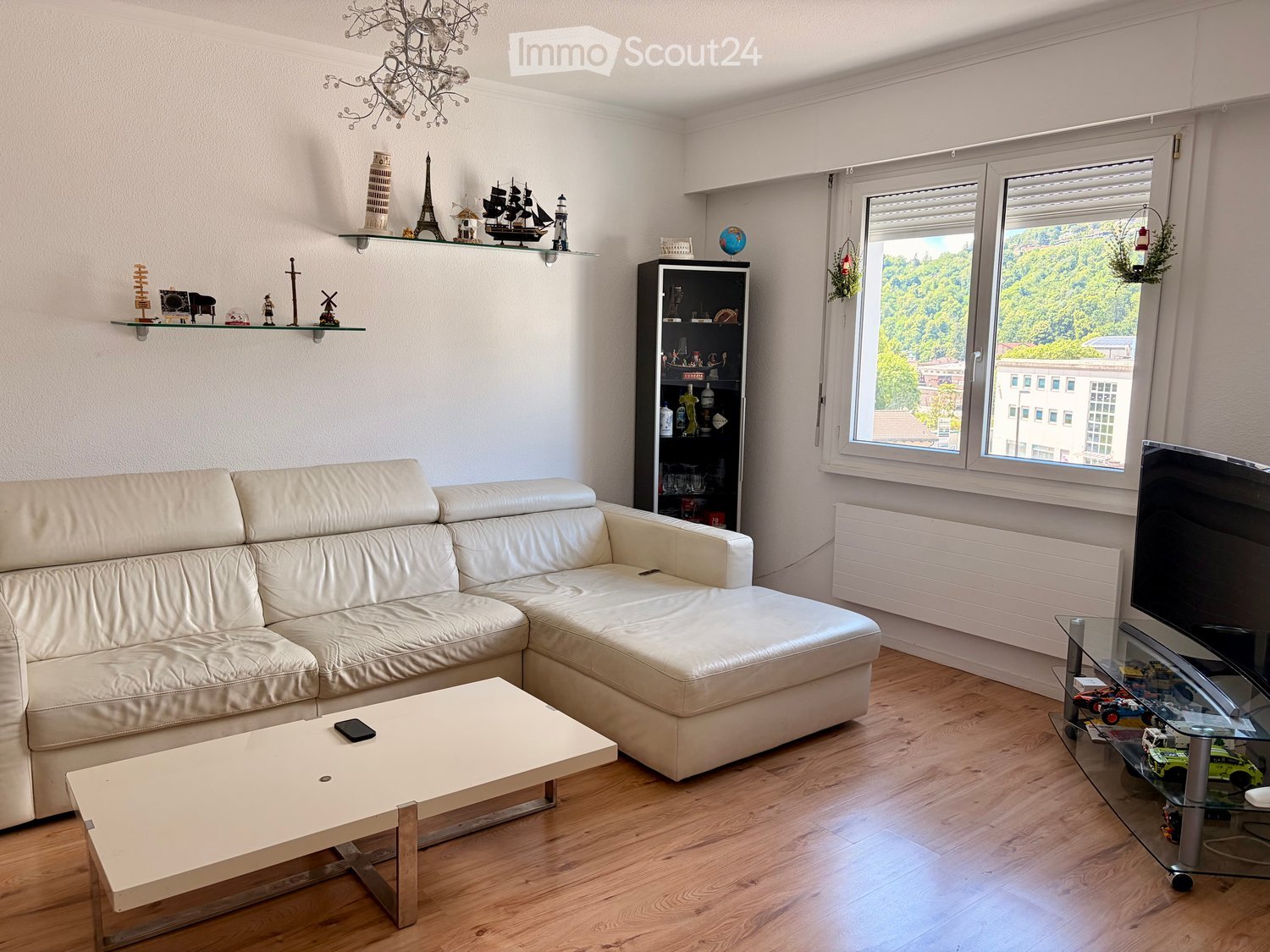 Modern living room with a white leather sofa, a light-colored coffee table, a TV stand with a flat-screen TV, a glass shelf with decorative items, and a chandelier.