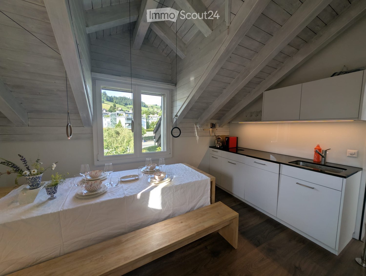 Bright, airy room with exposed wooden beams, a dining table set for four, a modern kitchen area with white cabinets and black countertops, and a view of greenery through large windows.