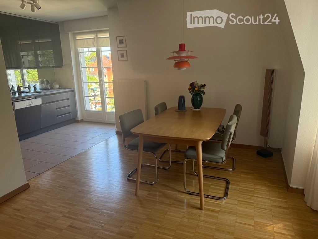 A modern dining area with a wooden dining table and four chairs, a vase with flowers on the table, and a sleek kitchen area with dark cabinets, a sink, and a dishwasher. The room features wooden flooring and large windows with blinds.