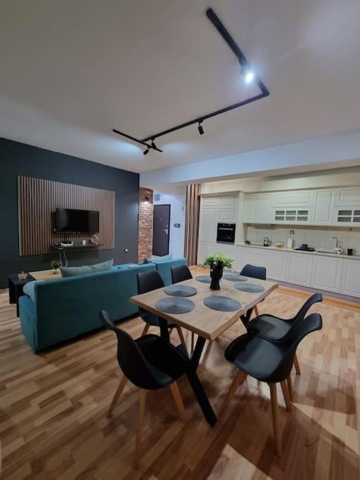A modern, open-plan living and dining area with a blue sofa, a wooden dining table with black chairs, a television mounted on a dark wall, and a white kitchen with cabinets, an oven, and a sink.