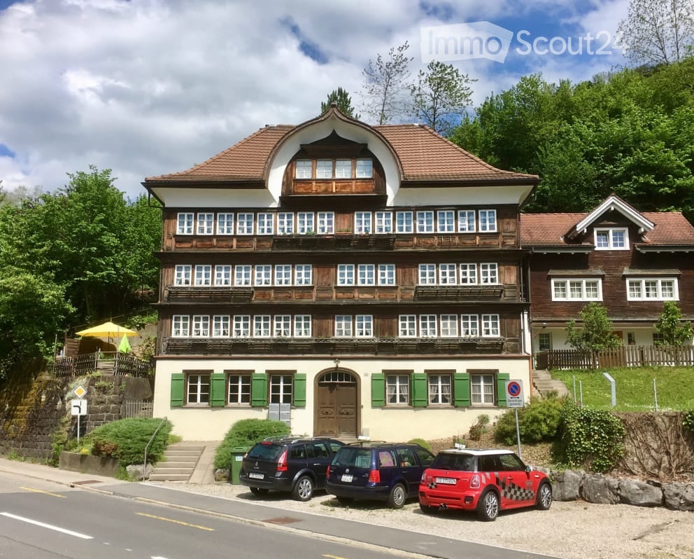A three-story traditional house with a wooden facade and green shutters, featuring multiple windows and a central entrance. The building is surrounded by greenery and has several parked cars in front.