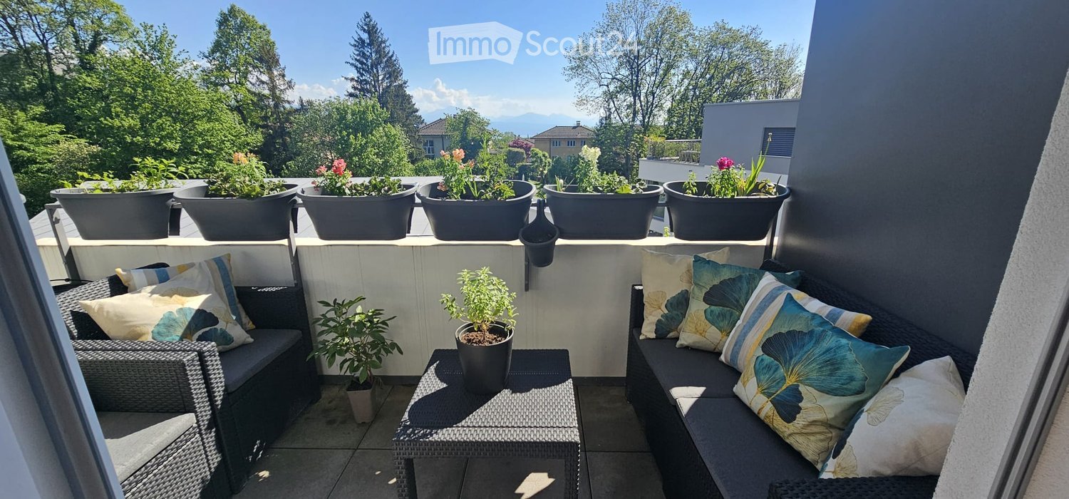 Balcony with a sofa, table, chairs, and potted plants, overlooking a green landscape with trees and mountains.