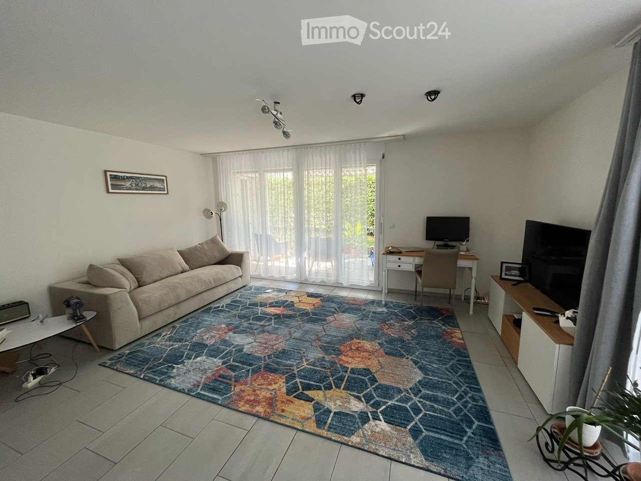Bright living room with a beige sofa, colorful geometric rug, coffee table, desk with chair, television, and floor lamp. Large windows with sheer curtains let in natural light.