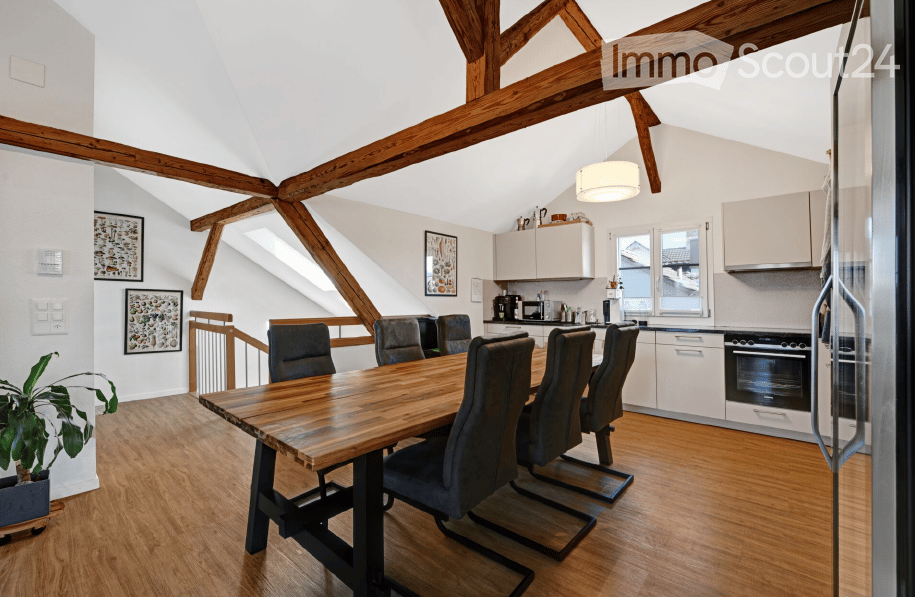 Open-plan dining and kitchen area with exposed wooden beams, a large wooden dining table surrounded by six chairs, modern kitchen cabinets, and a coffee machine on the counter.
