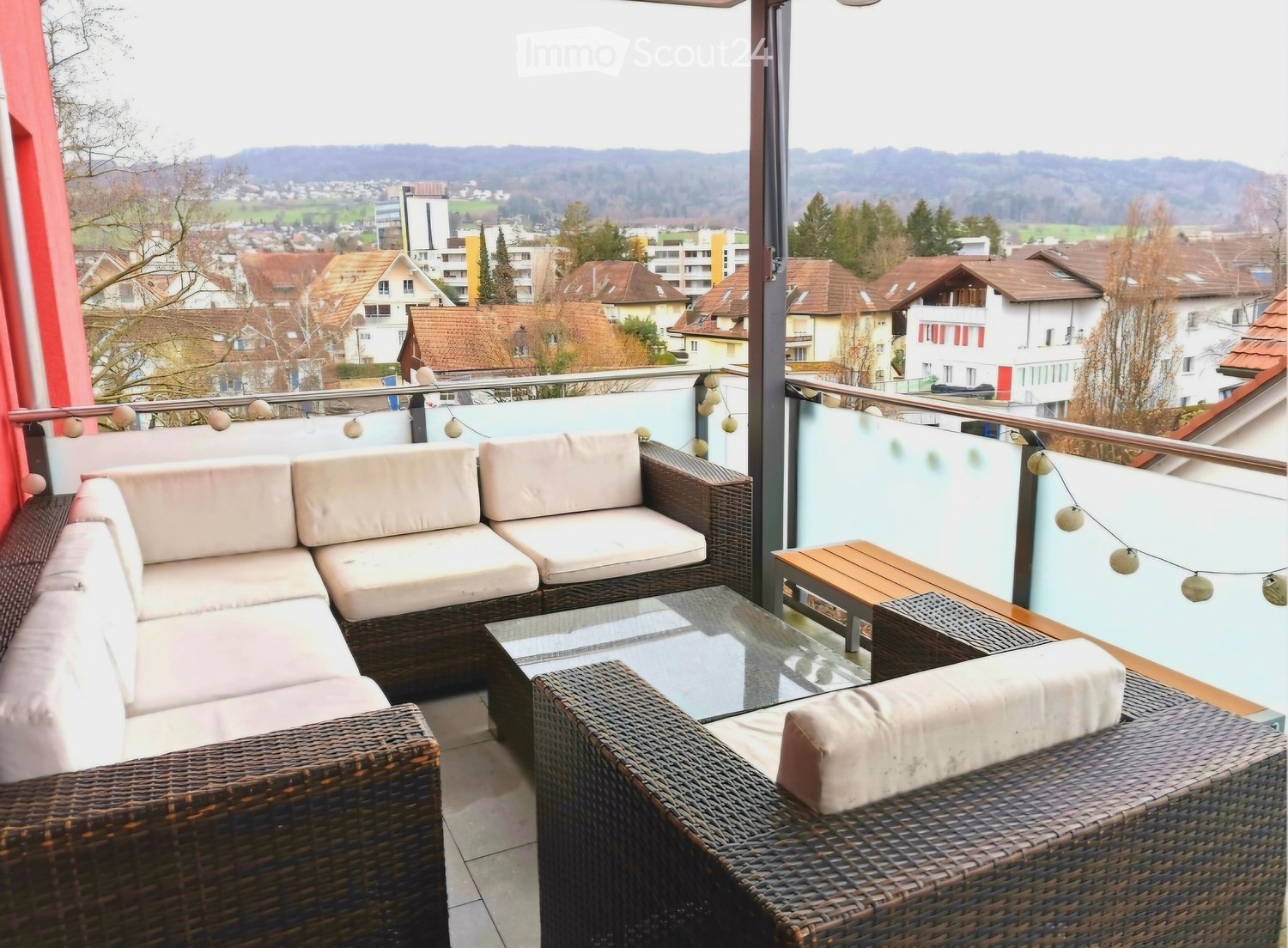 Balcony with wicker furniture, glass table, and scenic city view.