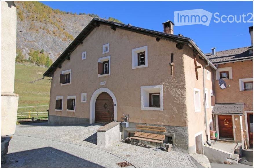 A rustic, two-story building with a beige facade, multiple windows, and a wooden door. A wooden bench is placed near the entrance, and the building is situated in a scenic, mountainous area with a clear blue sky.