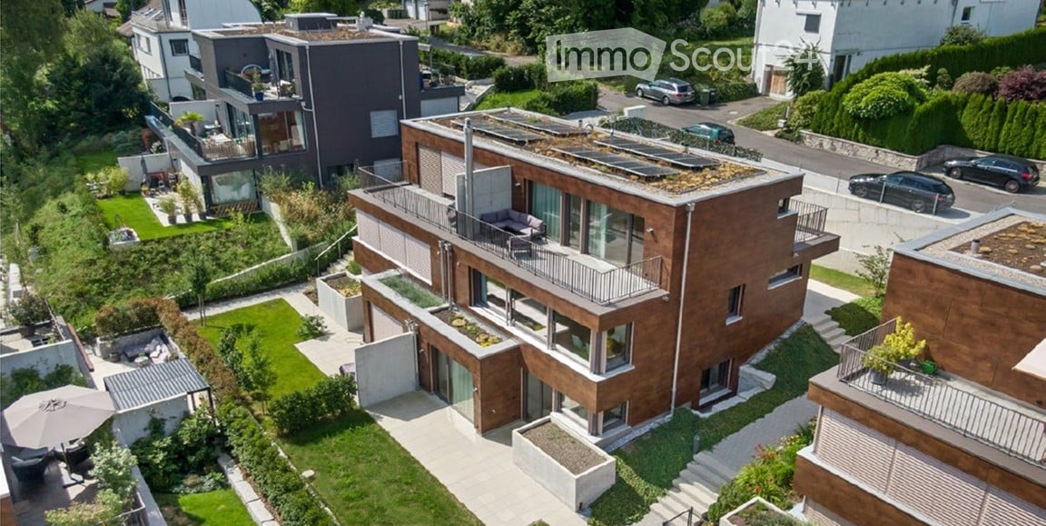 Aerial view of a modern, multi-story residential building with a green roof, balconies, and surrounding landscaped gardens. The building features contemporary architecture with large windows and a mix of wood and concrete materials.