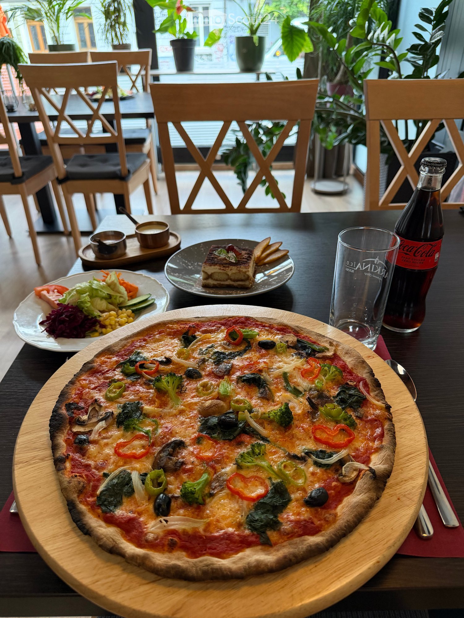 A dining table set with a pizza, salad, and other dishes, accompanied by a bottle of Coca-Cola and a glass, with wooden chairs and large windows in the background.