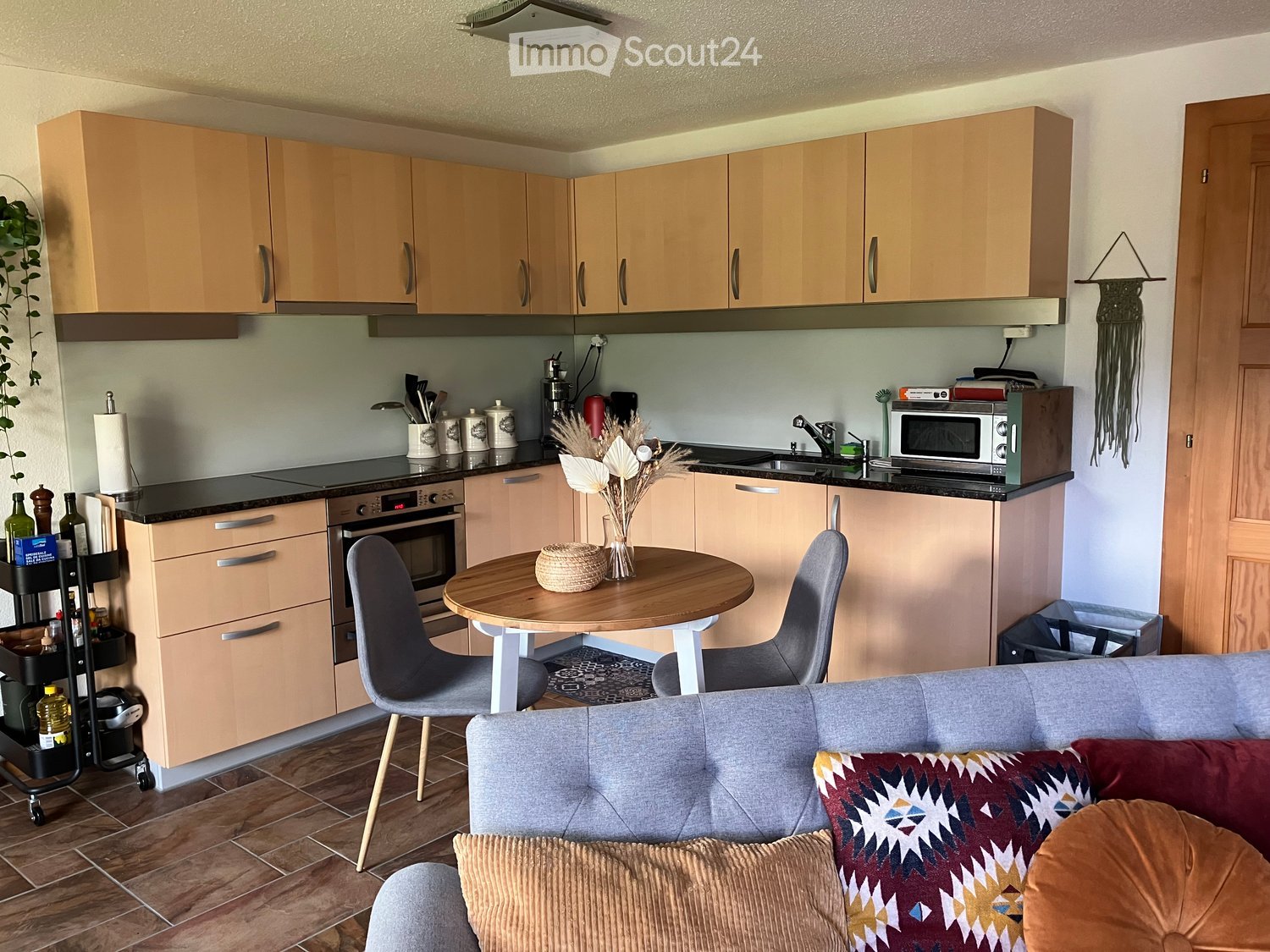 A modern, open-plan living and dining area with a grey sofa, patterned cushions, a round wooden dining table with two chairs, a kitchen area with light wood cabinets, an oven, microwave, and a sink.