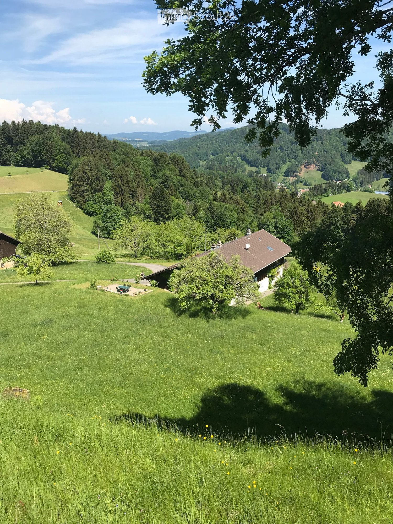 A peaceful countryside scene with a small house, lush green fields, and distant forested hills under a clear blue sky.