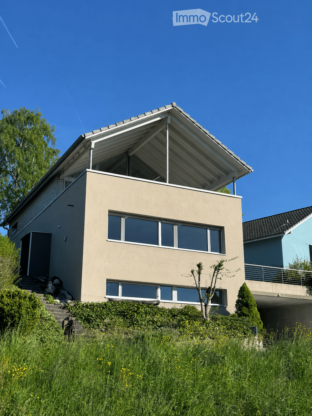 Modern two-story house with a flat roof and large windows, situated on a raised ground floor with a grassy slope leading up to it.
