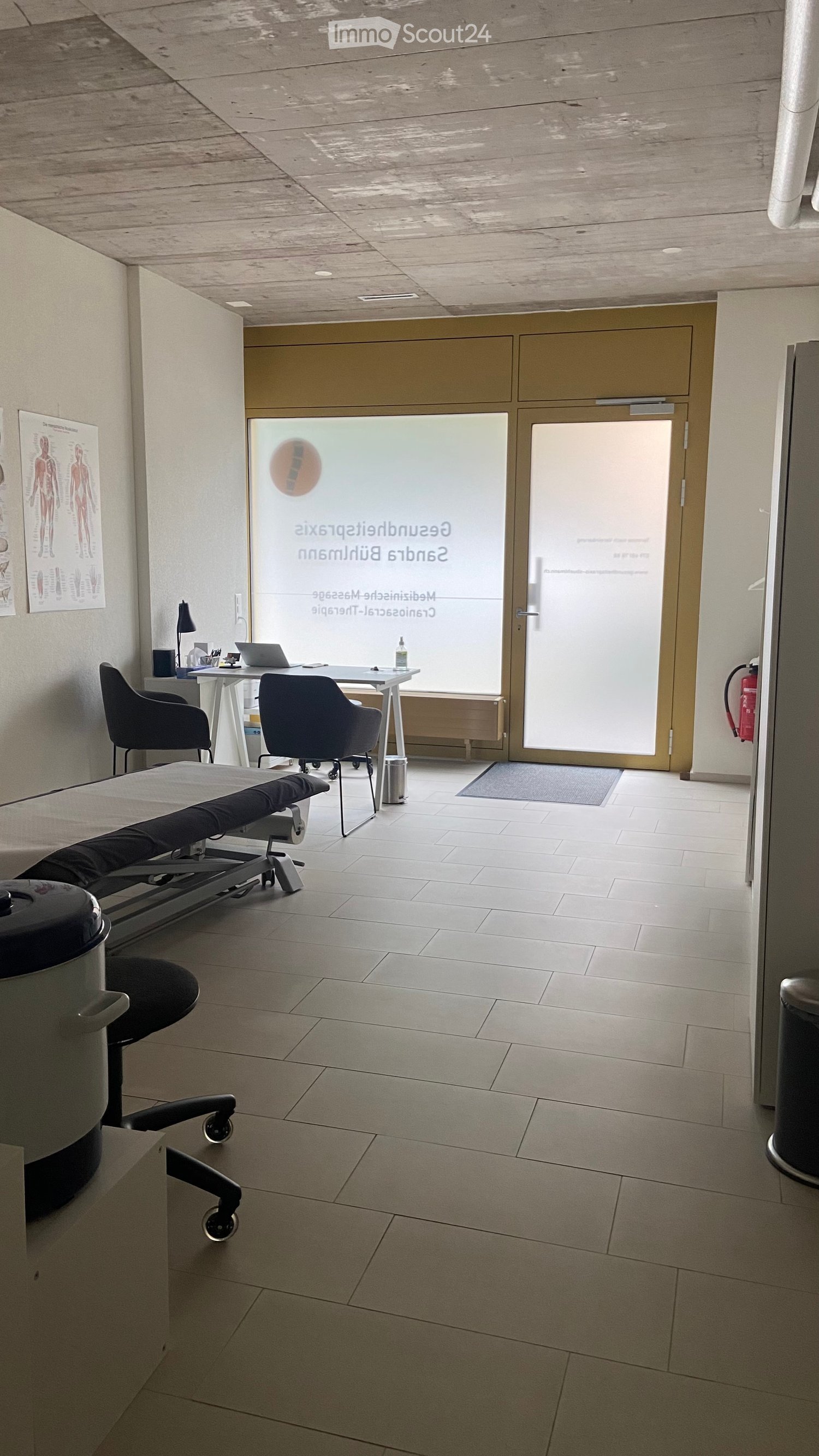 Modern medical or therapy room with tiled floor, large glass door, examination table, chairs, desk with laptop, and a lamp.