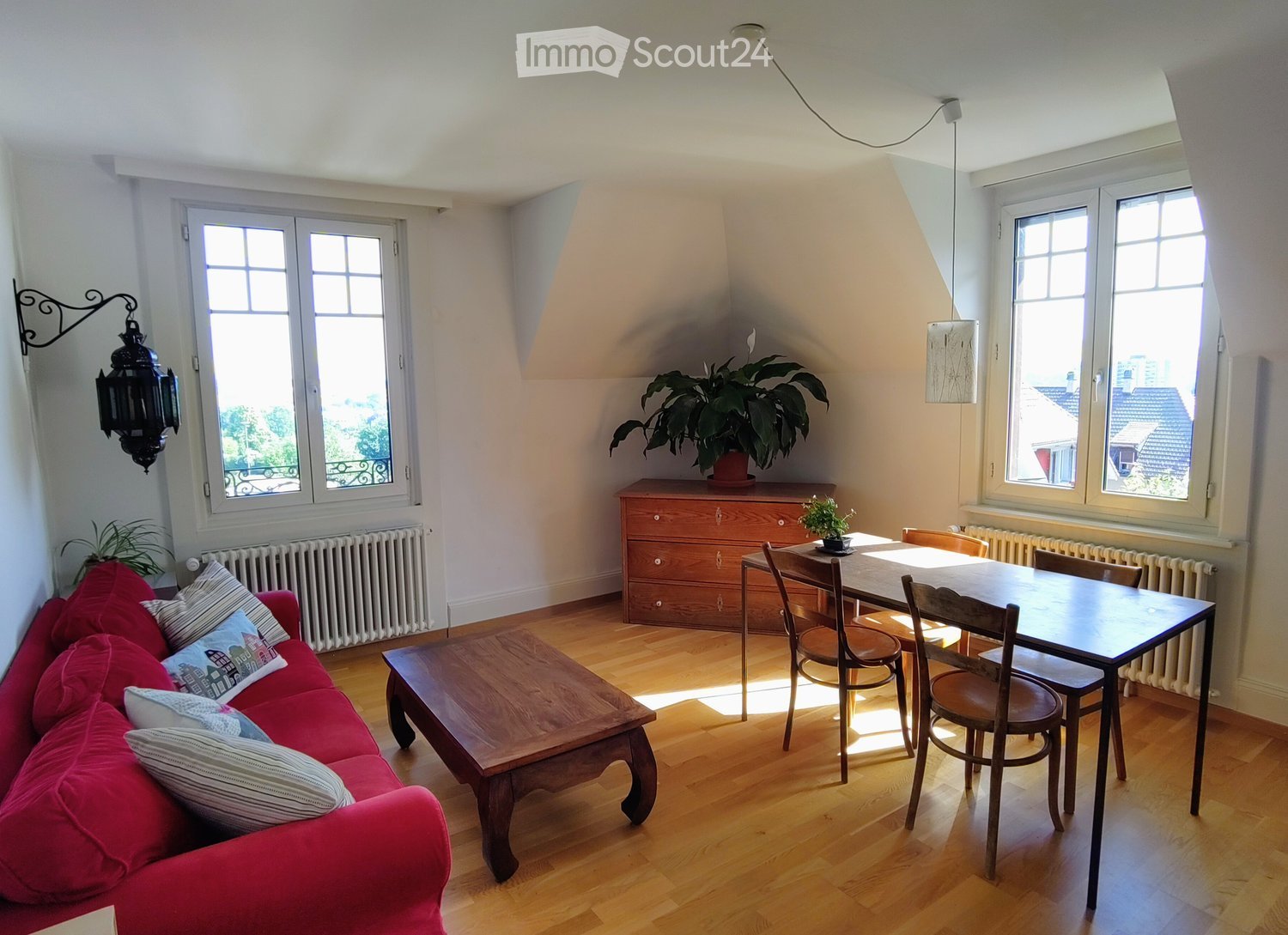 Bright living room with wooden flooring, red sofa, coffee table, dining table, chairs, and a potted plant on a wooden cabinet.