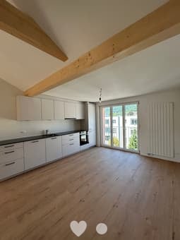 Bright, open kitchen with white cabinets, dark countertops, and large windows offering a view. The floor is made of light wood, and the space is completely empty with no furniture or appliances.