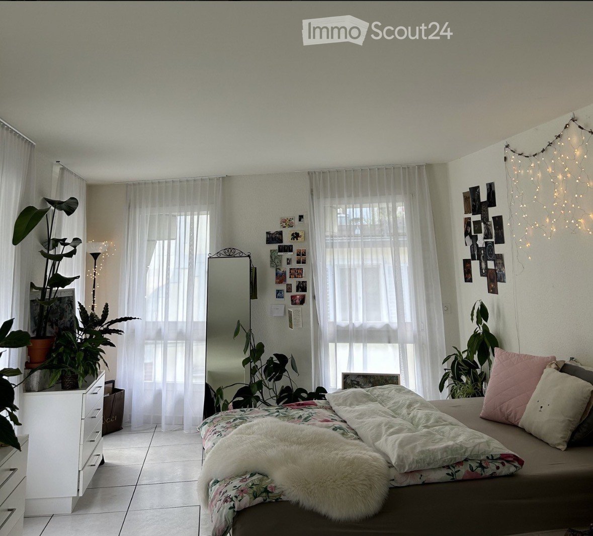 Cozy bedroom with a bed, floral duvet, fur throw, pink pillows, a mirror, a small dresser, and several potted plants.