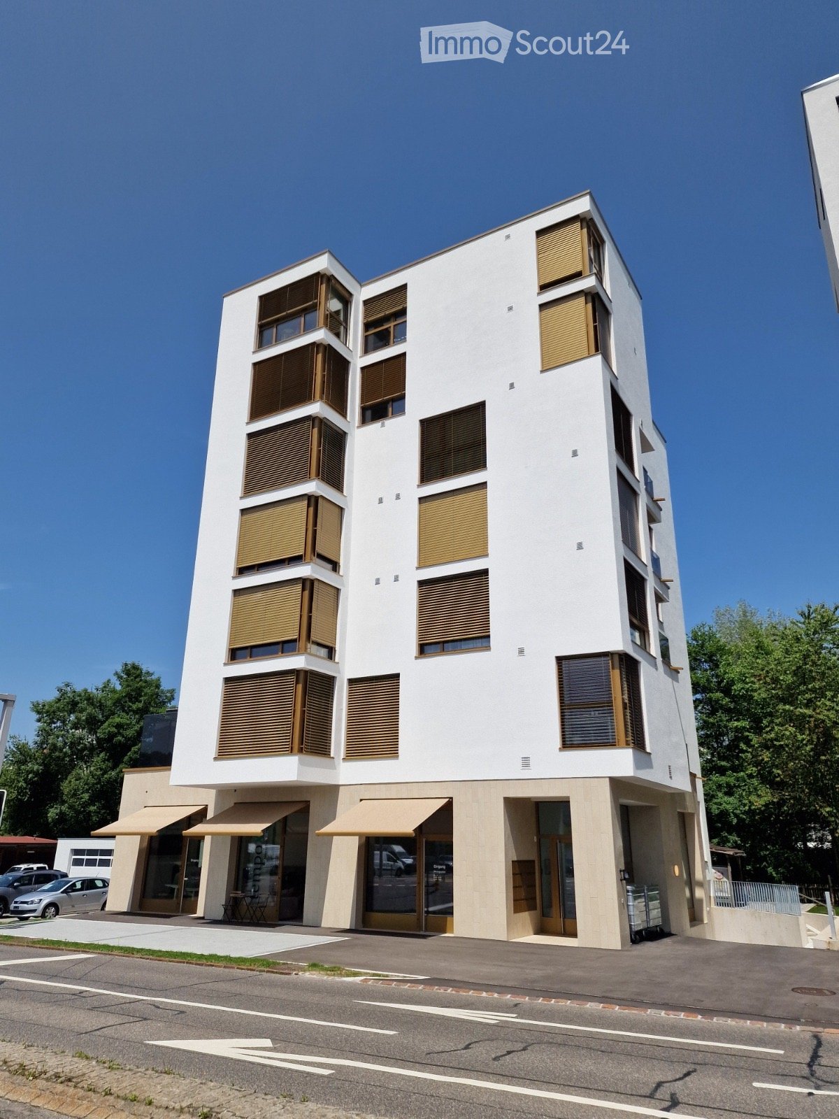 Modern multi-story building with white facade, large windows with wooden blinds, and a commercial storefront at the ground level.