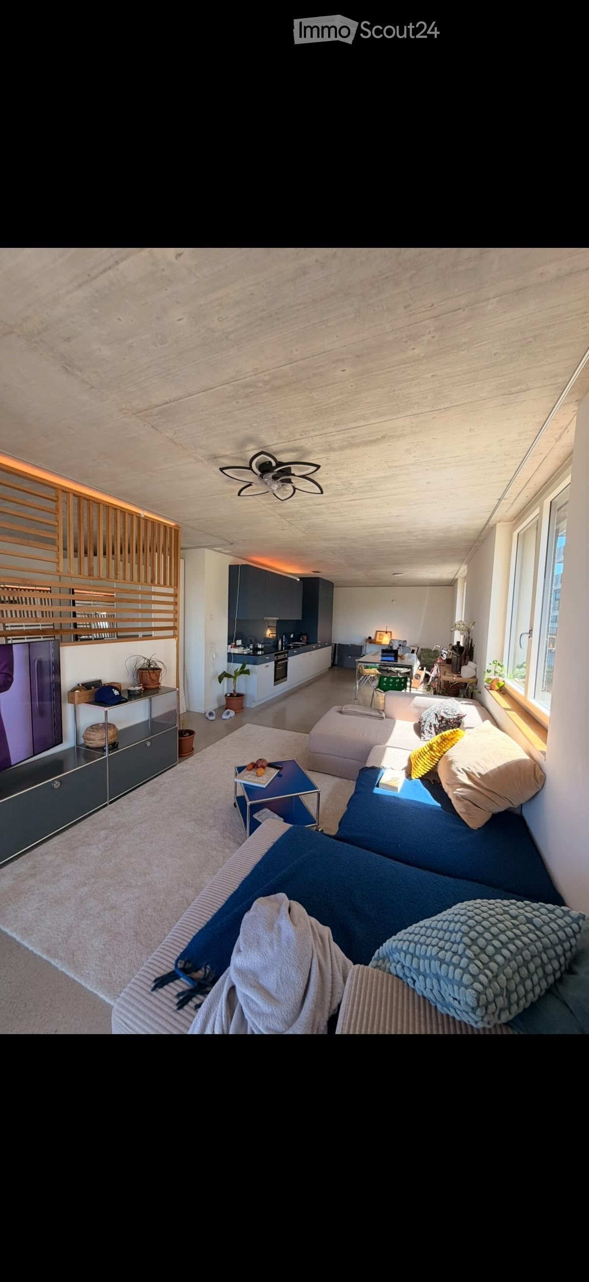 Modern open-plan living and dining area with a sectional sofa, coffee table, TV, and a small kitchen area with cabinets and appliances.