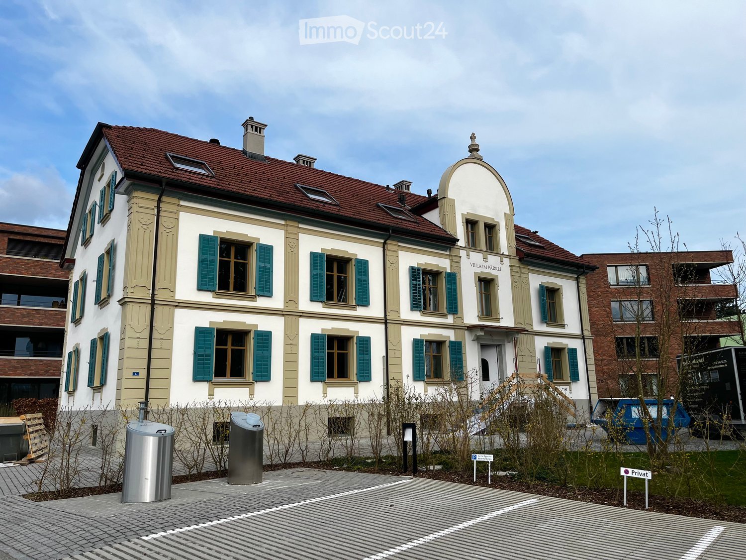 A two-story residential building with a red-tiled roof, white and beige facade, green shutters, and multiple windows. The building features a small dome-shaped turret and is surrounded by a paved area with trash bins and a sign indicating private property