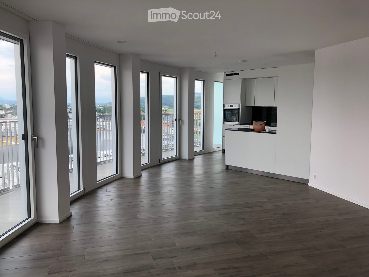 Modern, open-plan living and kitchen area with dark wooden flooring, white cabinetry, and large glass doors leading to a balcony with scenic views.