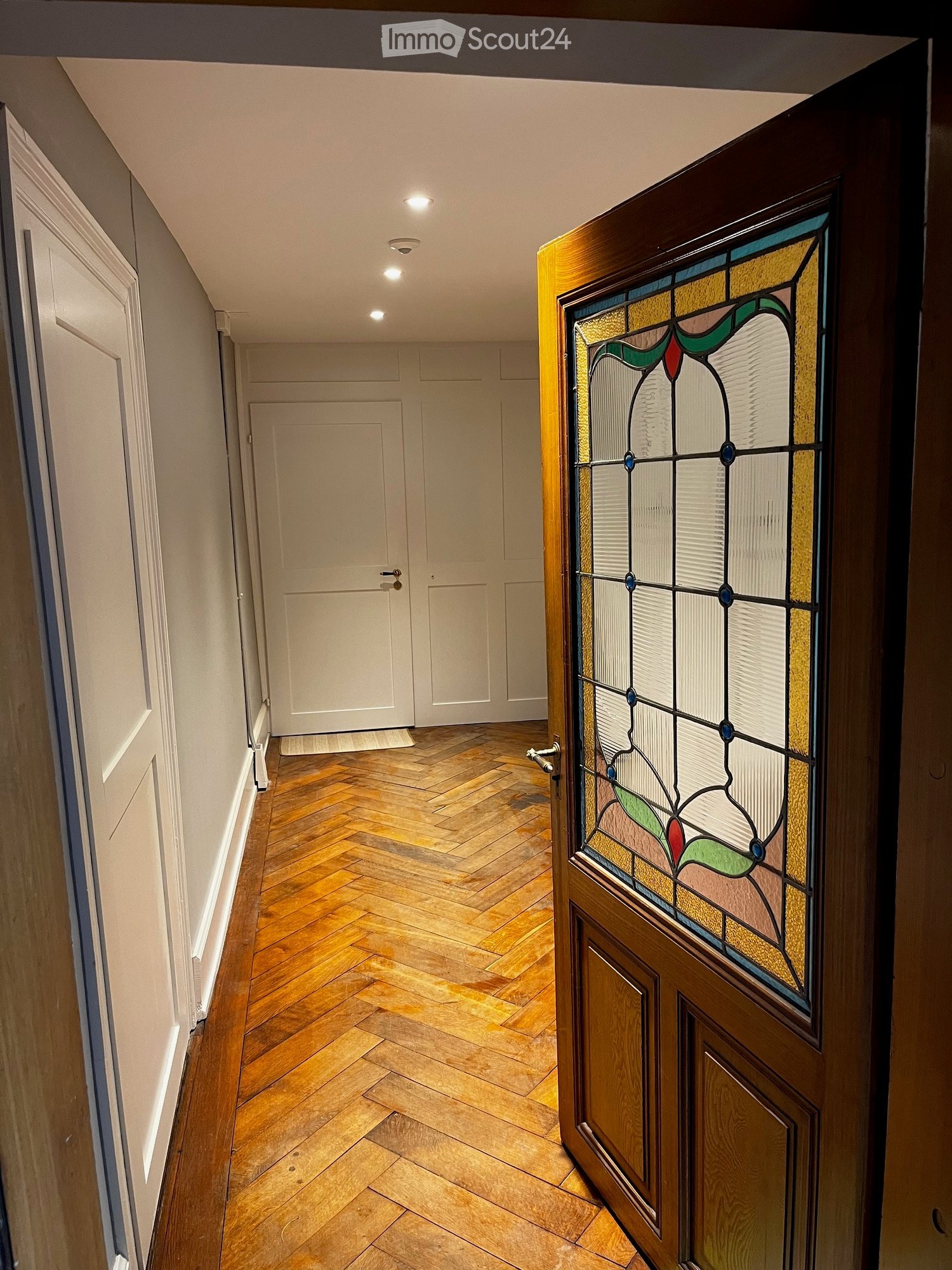 A narrow hallway with herringbone wooden flooring, white walls, and several closed doors, including one with stained glass.
