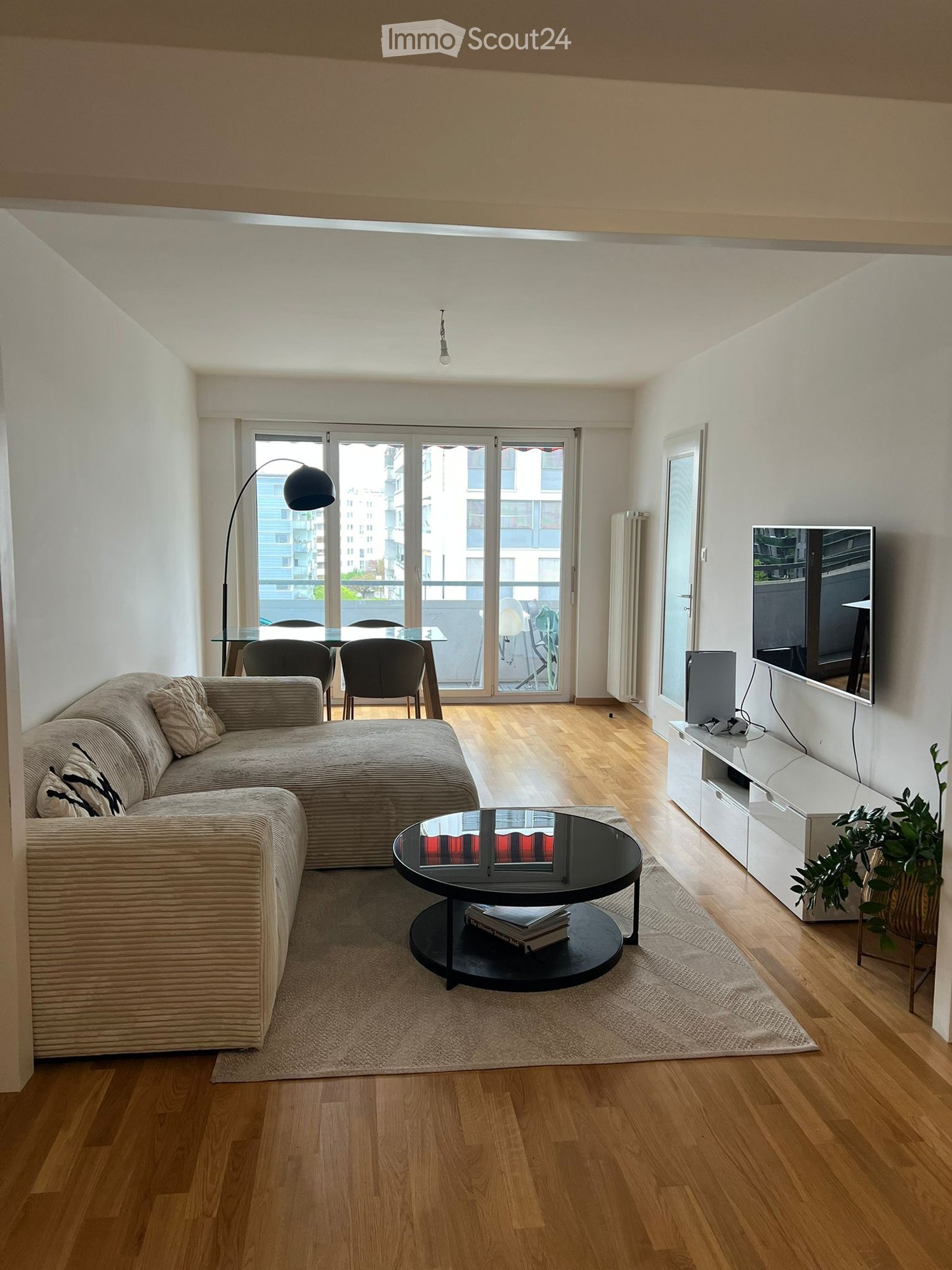modern living room with a large sofa, coffee table, TV, dining table, chairs, floor lamp, wooden floor, white walls, balcony with a view