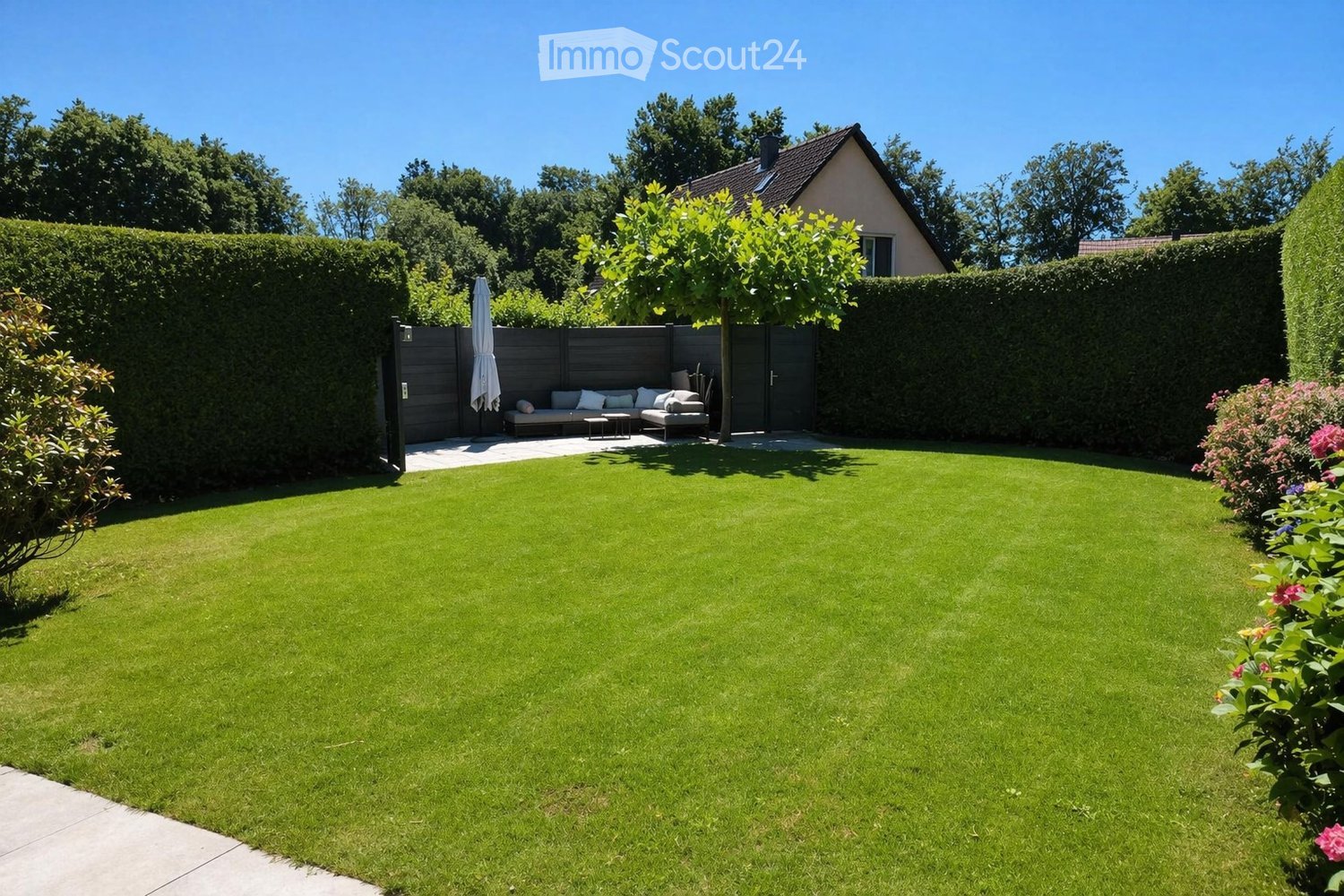 Well-maintained garden with a neatly trimmed lawn, a small seating area with a sofa and table, and a closed gate leading to the house.