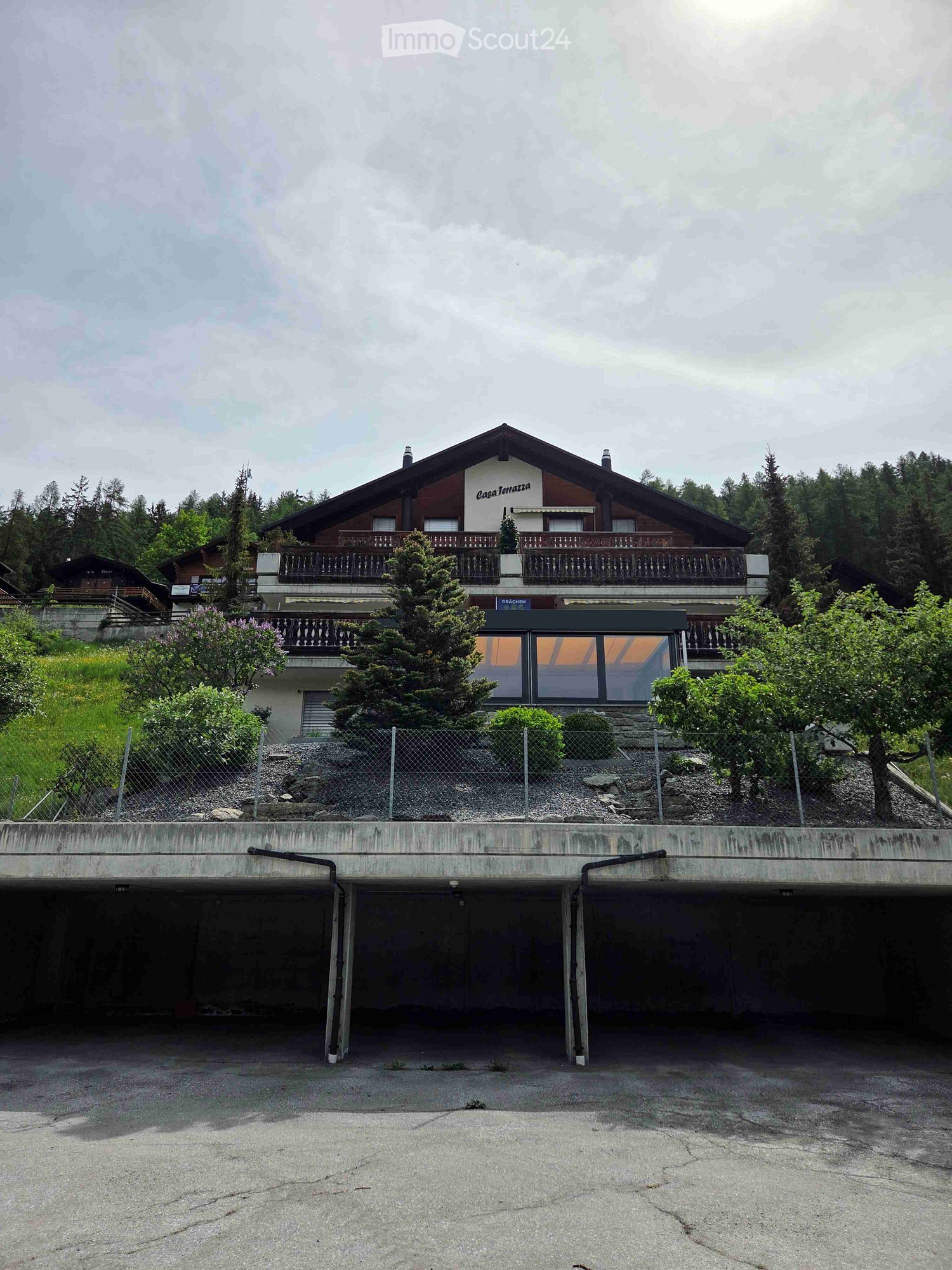 Two-story building with a balcony and garage underneath, surrounded by greenery and trees.