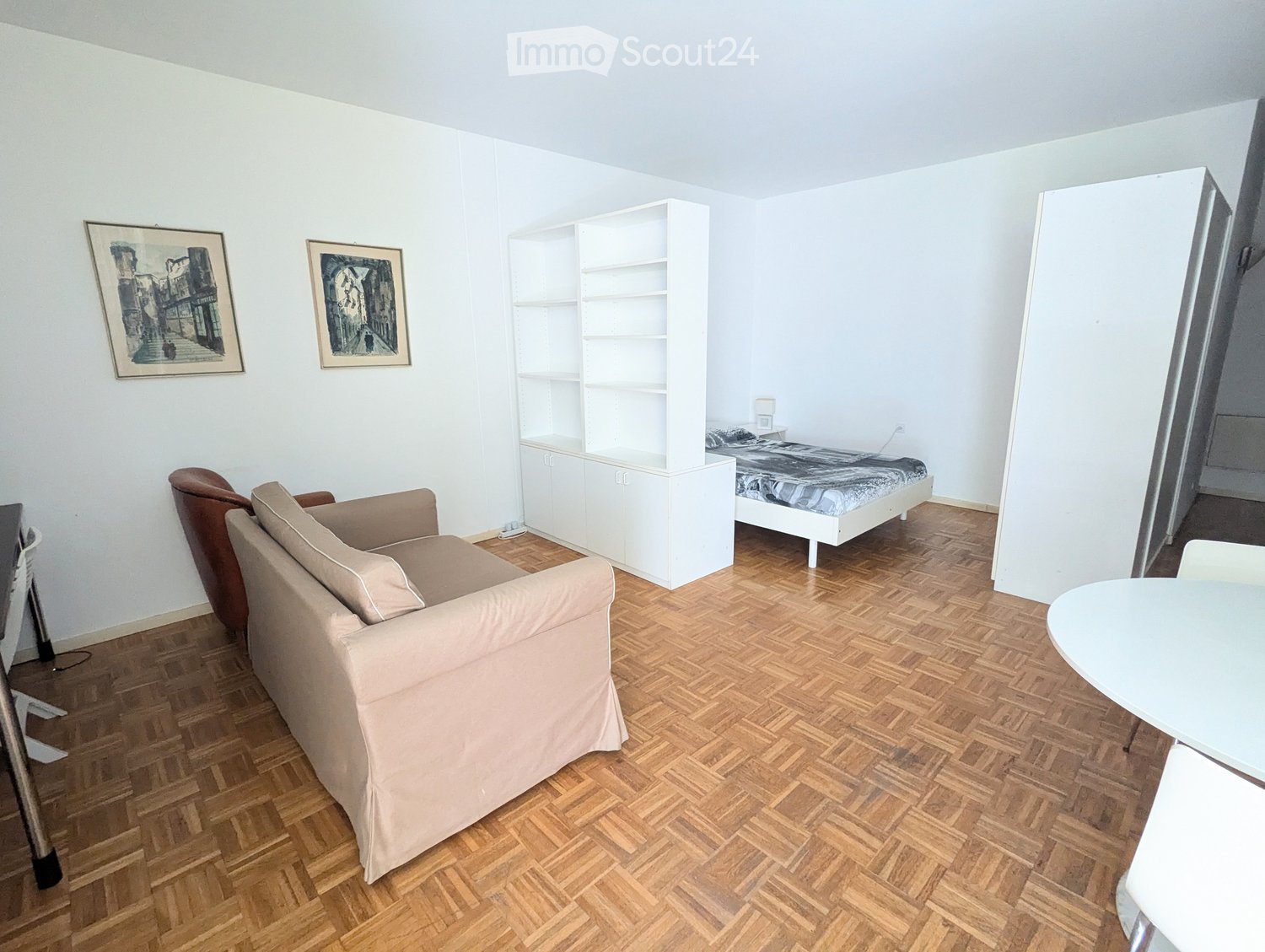 A furnished room with a sofa, armchair, small table, bed, white shelving unit, and two picture frames on the wall.