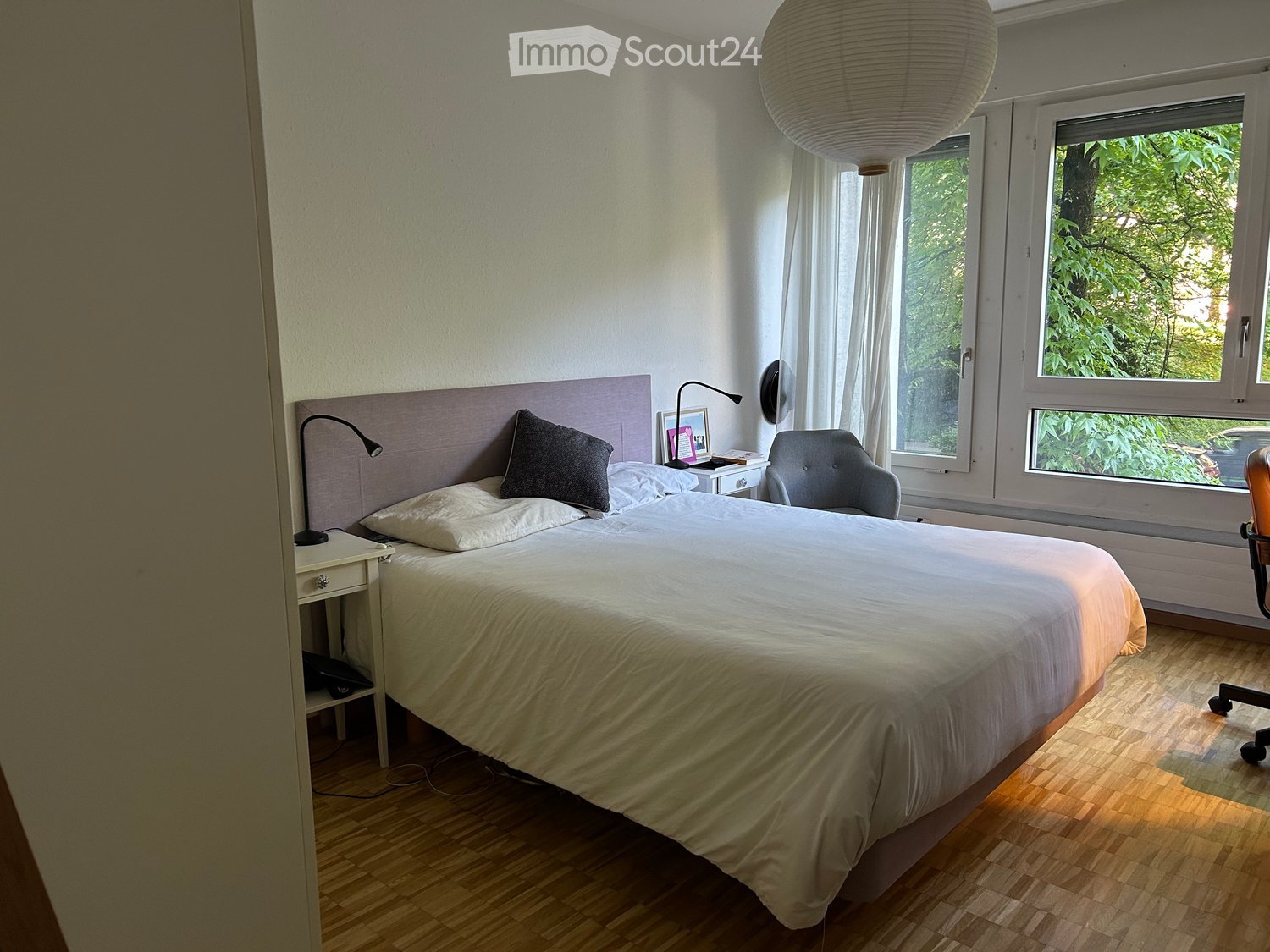 Cozy bedroom with a large bed, nightstand, chair, and a hanging paper lantern light fixture. The room features wooden flooring and large windows with white curtains.