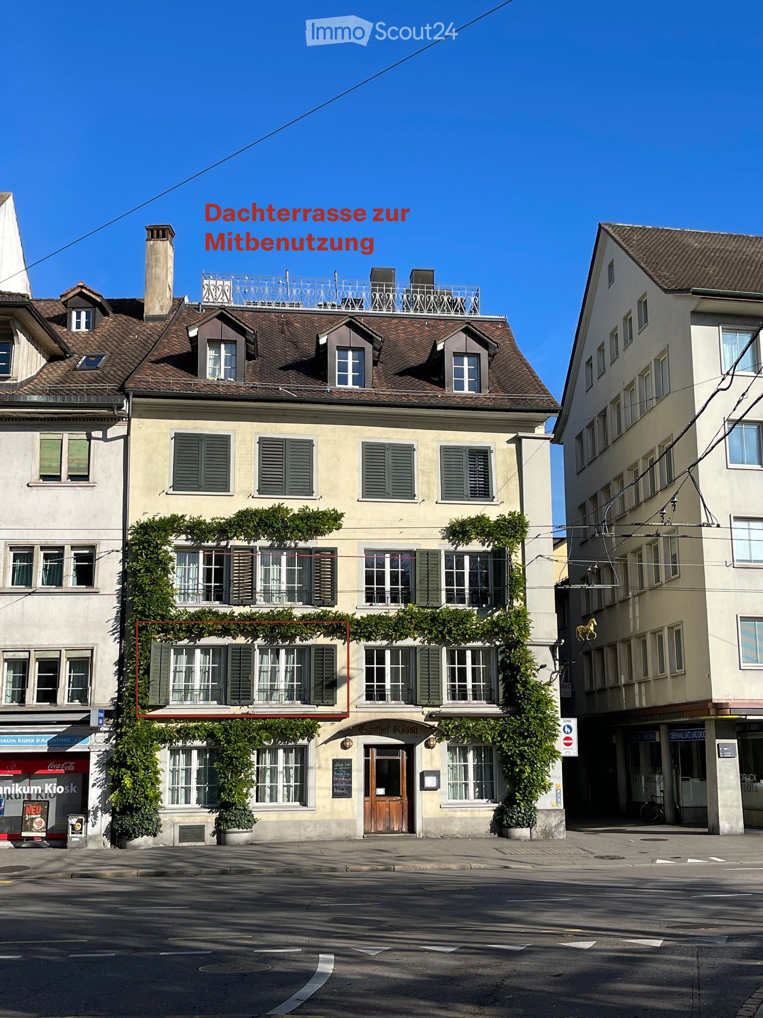 A multi-story building with a brown tiled roof, green shutters, and a balcony on the roof. The facade is covered with climbing plants, and there is a sign indicating a shared rooftop terrace.