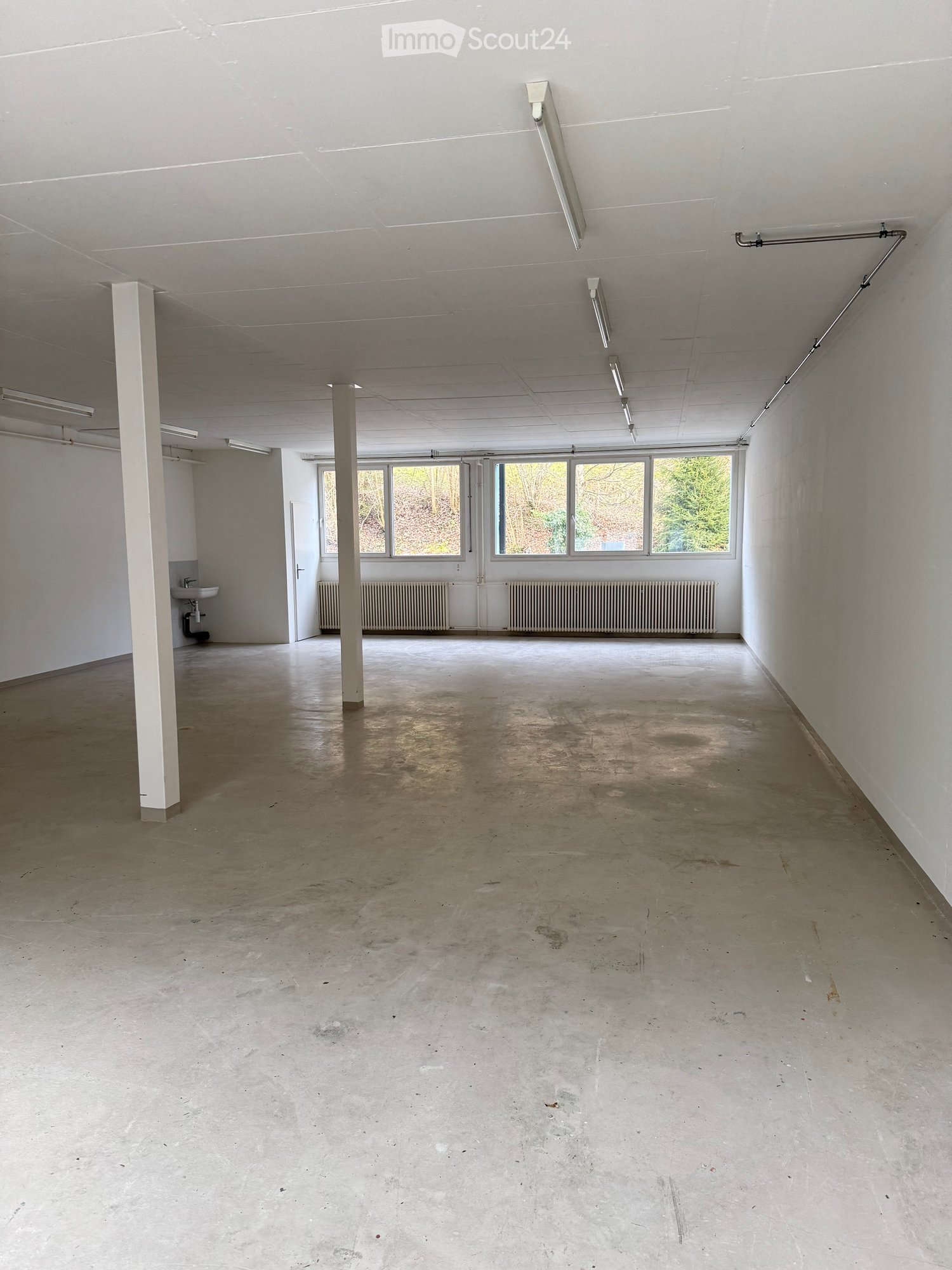 Empty room with concrete floor, white walls, and large windows. Features include a sink, radiator, and fluorescent lighting.