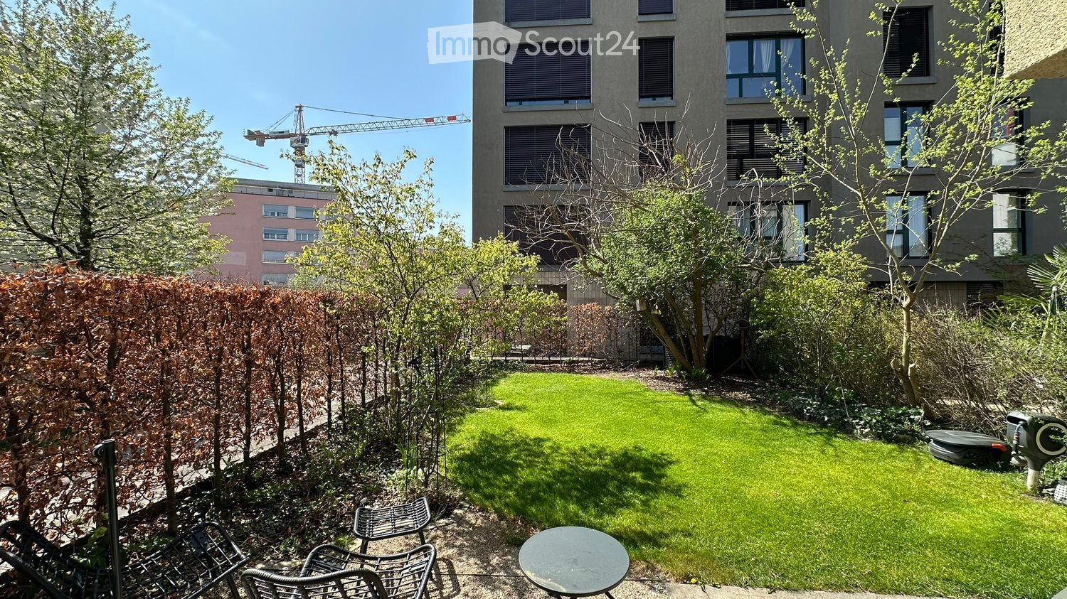 small garden with green grass, trees, shrubs, chairs, table, and a building in the background